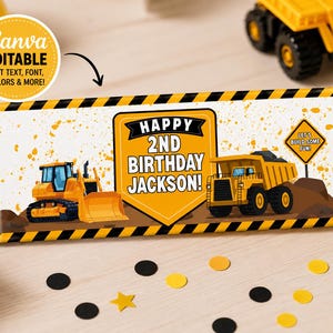 May include: A rectangular candy bar wrapper featuring a construction theme with yellow and black hazard stripes. It displays a yellow bulldozer, a yellow dump truck, and text that reads: HAPPY 2ND BIRTHDAY JACKSON! and LET'S BUILD SOME FUN!