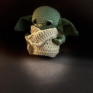 K&ouml;nnte beinhalten: Eine kleine, handgefertigte H&auml;kelpuppe mit einem gr&uuml;nen, runden Kopf, gro&szlig;en, dunklen, gl&auml;nzenden Augen und breiten, spitzen Ohren. Die Figur ist in ein strukturiertes, beigefarbenes Kleidungsst&uuml;ck geh&uuml;llt, das den K&ouml;rper bedeckt und die Arme zeigt.