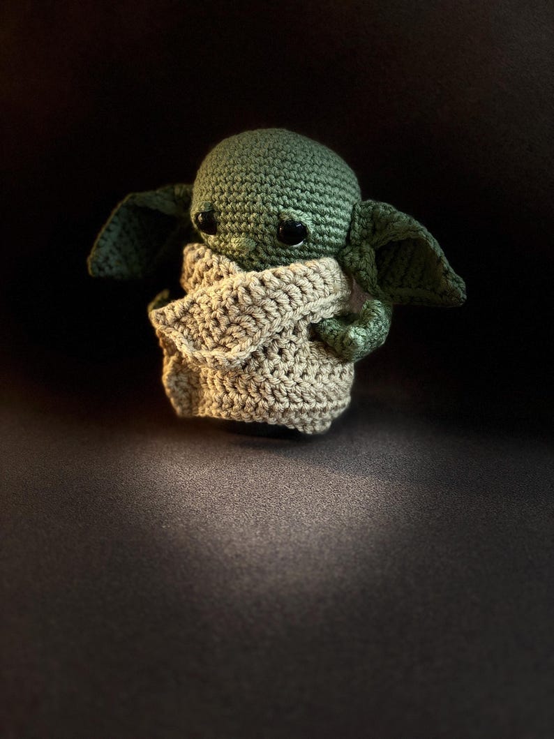 K&ouml;nnte beinhalten: Eine kleine, handgefertigte H&auml;kelpuppe mit einem gr&uuml;nen, runden Kopf, gro&szlig;en, dunklen, gl&auml;nzenden Augen und breiten, spitzen Ohren. Die Figur ist in ein strukturiertes, beigefarbenes Kleidungsst&uuml;ck geh&uuml;llt, das den K&ouml;rper bedeckt und die Arme zeigt.