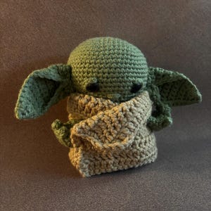 K&ouml;nnte beinhalten: Eine kleine, handgefertigte H&auml;kelpuppe mit einem runden gr&uuml;nen Kopf, gro&szlig;en spitzen Ohren und schwarzen Perlenaugen. Die Figur ist in ein strukturiertes, hellbeiges H&auml;kelgewand geh&uuml;llt, das den K&ouml;rper bedeckt und die gr&uuml;nen Arme teilweise freil&auml;sst.