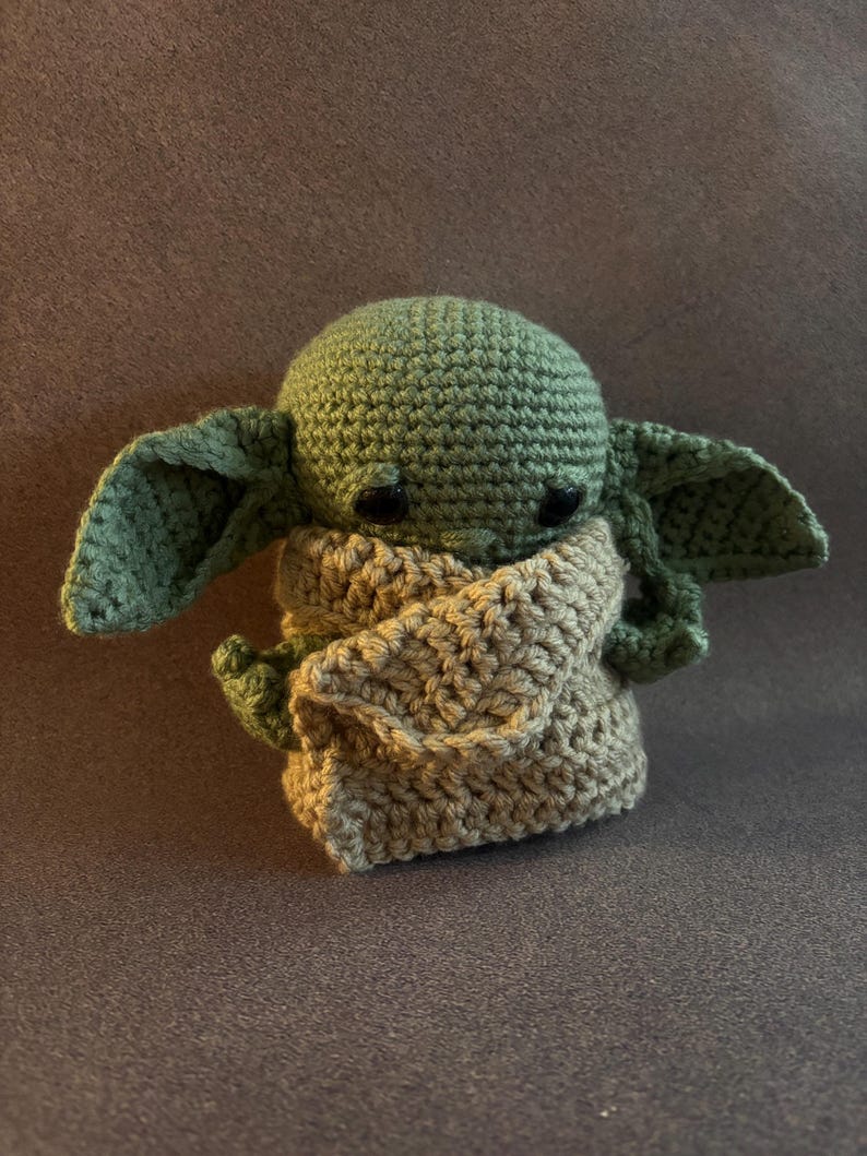 K&ouml;nnte beinhalten: Eine kleine, handgefertigte H&auml;kelpuppe mit einem runden gr&uuml;nen Kopf, gro&szlig;en spitzen Ohren und schwarzen Perlenaugen. Die Figur ist in ein strukturiertes, hellbeiges H&auml;kelgewand geh&uuml;llt, das den K&ouml;rper bedeckt und die gr&uuml;nen Arme teilweise freil&auml;sst.