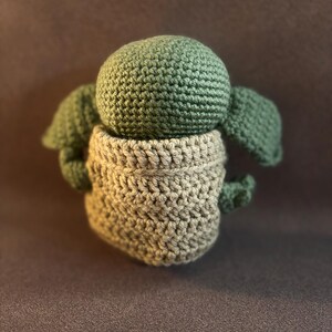 K&ouml;nnte beinhalten: Eine kleine, handgefertigte H&auml;kelpuppe mit einem runden, salbeigr&uuml;nen Kopf, gro&szlig;en abstehenden Ohren und einem strukturierten, beigefarbenen Kleidungsst&uuml;ck, das den Unterk&ouml;rper bedeckt. Die Figur ist mit sichtbaren Garnmaschen gearbeitet.