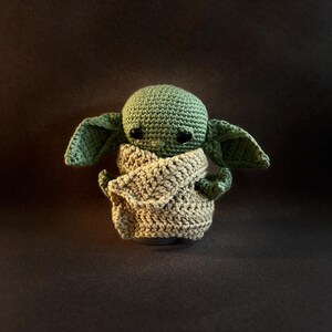 K&ouml;nnte beinhalten: Eine kleine, handgefertigte H&auml;kelpuppe mit einem runden gr&uuml;nen Kopf, gro&szlig;en, dunklen, gl&auml;nzenden Augen und markanten spitzen Ohren. Die Figur ist in ein strukturiertes, beigefarbenes Garnkleid geh&uuml;llt, das den Oberk&ouml;rper bedeckt und weich wirkt.