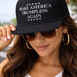 May include: Black baseball cap featuring the white text "MAKE AMERICA TRUMPLESS AGAIN". The cap has a flat peak and a structured crown. The text is centered on the front, above and below a blue line and stars.