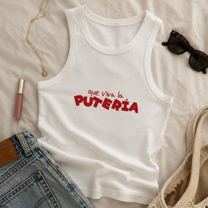 Puede incluir: Una camiseta de tirantes blanca de canalé con el texto rojo "que viva la PUTERÍA" impreso en el pecho. La prenda tiene un cuello redondo y tirantes anchos, presentada como una pieza de ropa independiente con un diseño limpio y sencillo.
