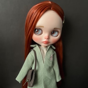 May include: A doll with long, red hair wearing a light green jacket with a brown purse. The doll has blue eyes and a light pink blush.
