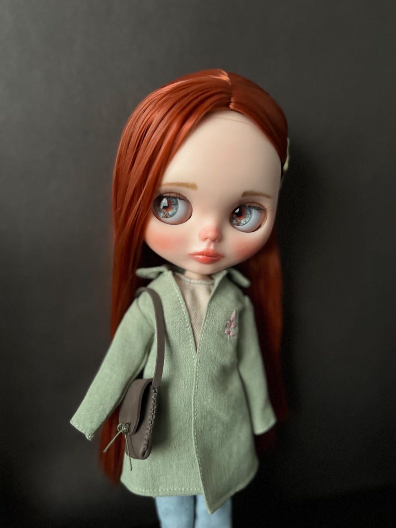 May include: A doll with long, red hair wearing a light green jacket with a brown purse. The doll has blue eyes and a light pink blush.