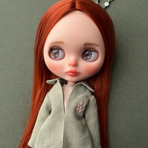 May include: A red-haired doll with blue eyes and a green coat. The doll is wearing a light green coat with a pink floral embroidery on the left side. The doll has a light green background.