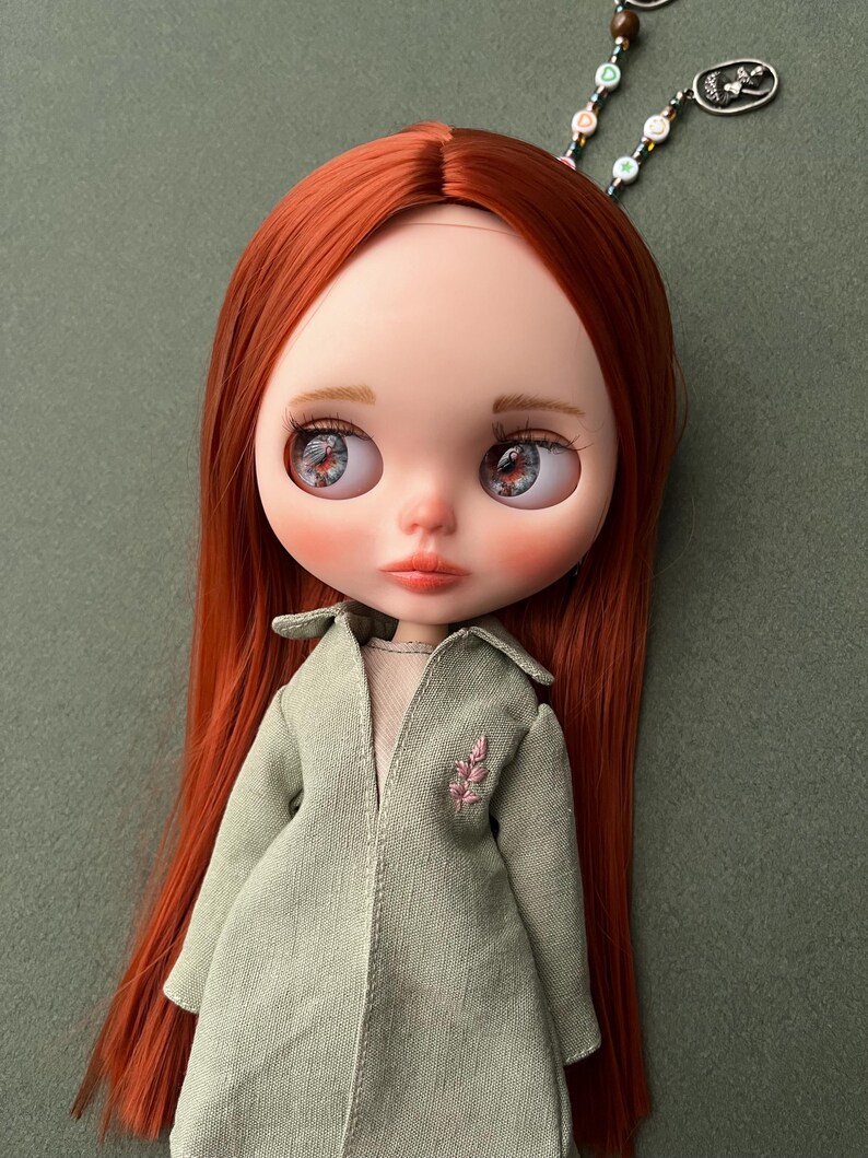 May include: A red-haired doll with blue eyes and a green coat. The doll is wearing a light green coat with a pink floral embroidery on the left side. The doll has a light green background.