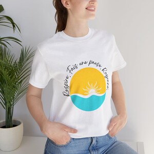 May include: White t-shirt with a circular design of a yellow sun, blue water, and a white sun. The French text "Respire. Fais une pause. Rayonne." is printed around the graphic in black. The shirt is made of a soft material.