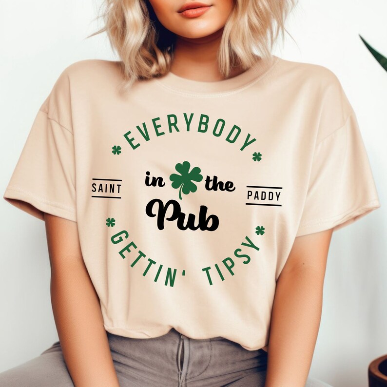Puede incluir: Una camiseta beige con un tr&eacute;bol verde y el texto "*EVERYBODY* in the Pub *GETTIN' TIPSY*" con las palabras "SAINT" y "PADDY" a cada lado del tr&eacute;bol.