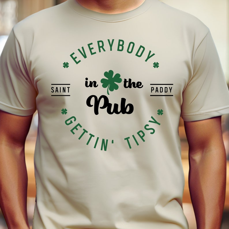 Puede incluir: Una camiseta beige con un tr&eacute;bol verde y el texto "Everybody in the Pub Gettin' Tipsy" en un c&iacute;rculo a su alrededor. Los textos "Saint" y "Paddy" est&aacute;n a ambos lados del tr&eacute;bol.