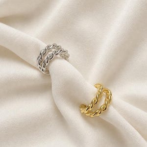 May include: Two ear cuffs, one in a polished silver tone and one in a polished gold tone. Each ear cuff features a double-band design with a twisted rope texture, creating a continuous circular shape with an open gap for placement.