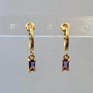 Könnte beinhalten: Ein Paar goldene Creolen mit einem baumelnden violetten Edelstein. Die Ohrringe befinden sich auf einem weißen Hintergrund.
