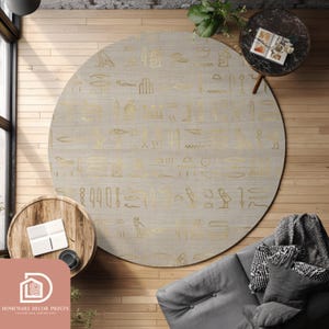 Könnte beinhalten: Ein großer, runder beigefarbener Teppich mit goldenen Hieroglyphenmustern. Der Teppich liegt auf einem Holzboden, umgeben von kleinen Tischen, einem grauen Sofa und einem Fenster. Das Design erinnert an ein altägyptisches Thema.