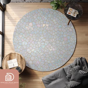 Op de afbeelding: Een ronde vloerkleed met een mozaïekpatroon in tinten wit, blauw en iriserende kleuren. Het kleed ligt op een houten vloer, met een bank en kleine tafels in de buurt. Het ontwerp van het kleed lijkt op een verzameling onregelmatige vormen.