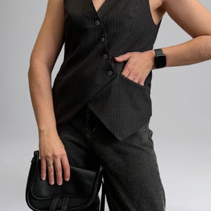 Pode incluir: Um colete listrado preto e branco com decote em V e bot&otilde;es. O colete &eacute; usado sobre uma camisa preta e cal&ccedil;as de ganga pretas. A pessoa est&aacute; usando um rel&oacute;gio preto no pulso esquerdo e uma bolsa de ombro de couro preta.