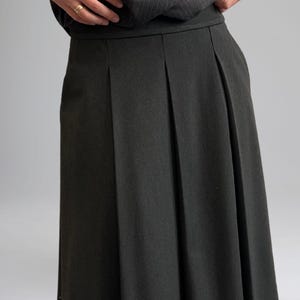 May include: A dark gray pleated skirt with a straight silhouette. The skirt is made of a textured fabric with a subtle pinstripe pattern.