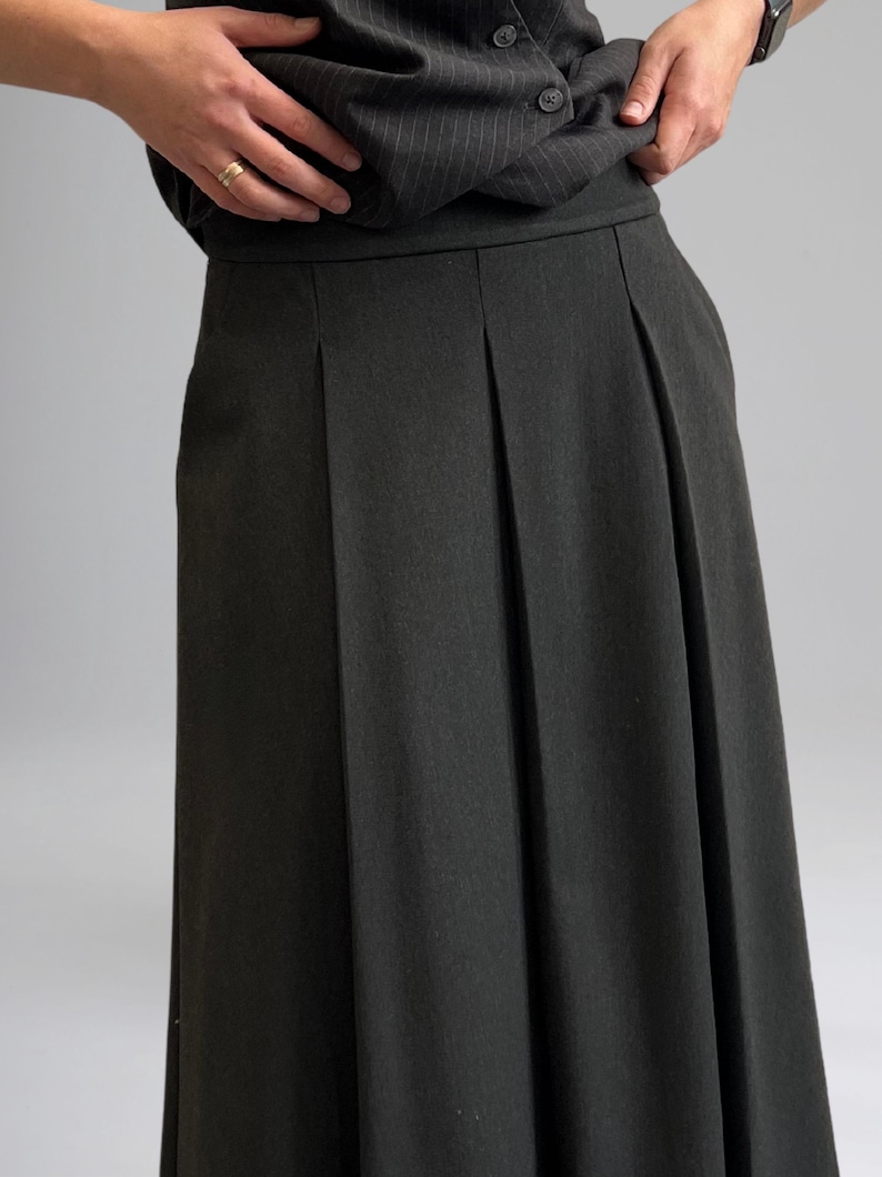 May include: A dark gray pleated skirt with a straight silhouette. The skirt is made of a textured fabric with a subtle pinstripe pattern.