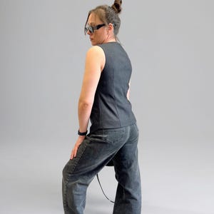 Pode incluir: Uma mulher vestindo um colete sem mangas listrado de preto e branco e jeans denim escuro. Ela est&aacute; usando &oacute;culos de sol e tem o cabelo preso em um coque.
