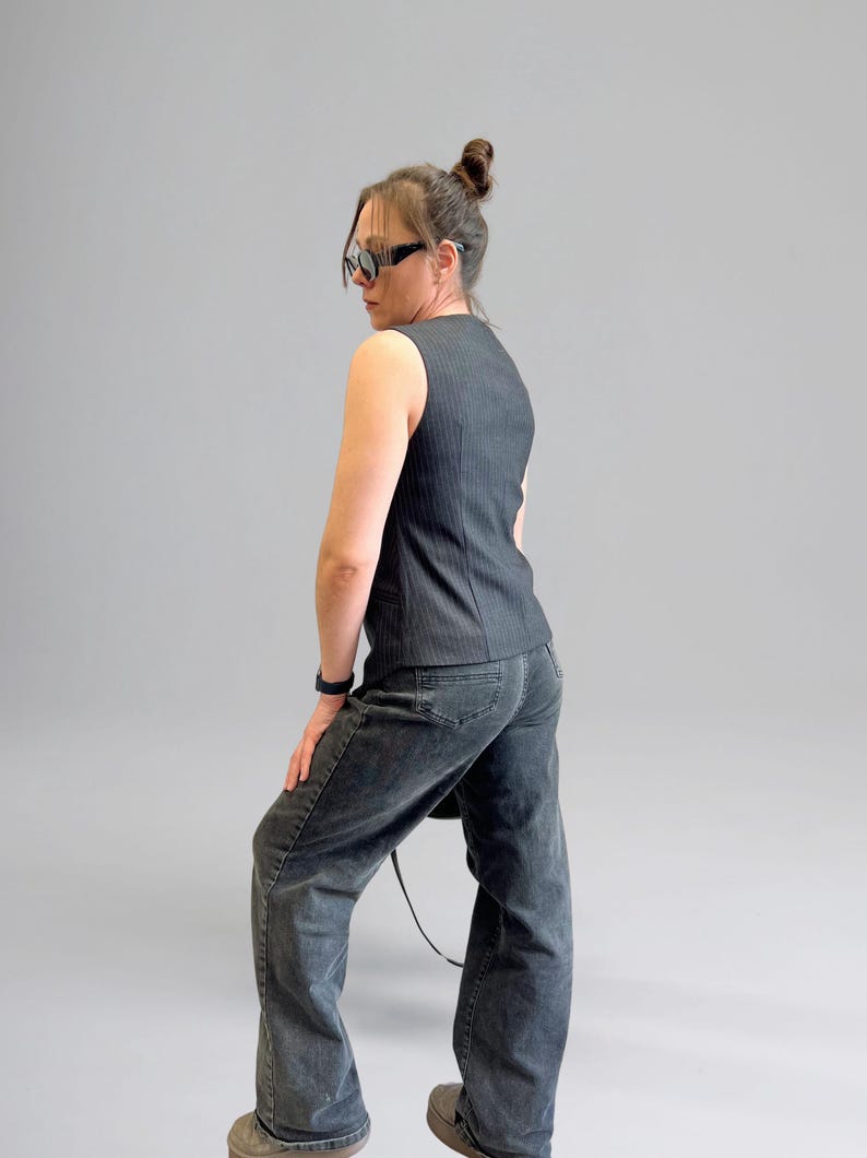 Pode incluir: Uma mulher vestindo um colete sem mangas listrado de preto e branco e jeans denim escuro. Ela est&aacute; usando &oacute;culos de sol e tem o cabelo preso em um coque.