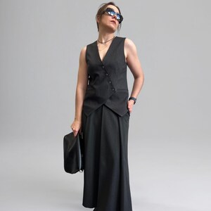 May include: A woman wearing a black and white striped vest with a black skirt and black boots. She is holding a black clutch bag and wearing sunglasses.