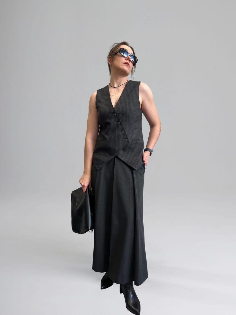 May include: A woman wearing a black and white striped vest with a black skirt and black boots. She is holding a black clutch bag and wearing sunglasses.