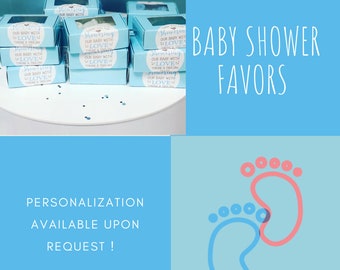 May include: Baby shower favors in light blue boxes with clear lids, stacked on a white surface. Each box has a sticker that reads "Showering our baby with love". The background is light blue with the text "Baby Shower Favors".
