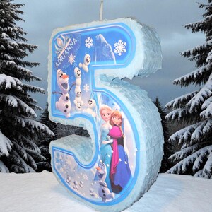 Puede incluir: Una pi&ntilde;ata grande con forma del n&uacute;mero 5, en azul claro y blanco, decorada con personajes de la pel&iacute;cula "Frozen". La pi&ntilde;ata tiene el nombre "Adrianna" impreso y est&aacute; decorada con copos de nieve y monta&ntilde;as nevadas.