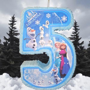 Puede incluir: Una pi&ntilde;ata grande, de color azul claro, con el n&uacute;mero 5 y personajes de la pel&iacute;cula Frozen. La pi&ntilde;ata tiene un borde azul claro e incluye im&aacute;genes de Olaf, Anna, Elsa y mu&ntilde;ecos de nieve. El nombre "Adrianna" est&aacute; escrito en una pancarta.