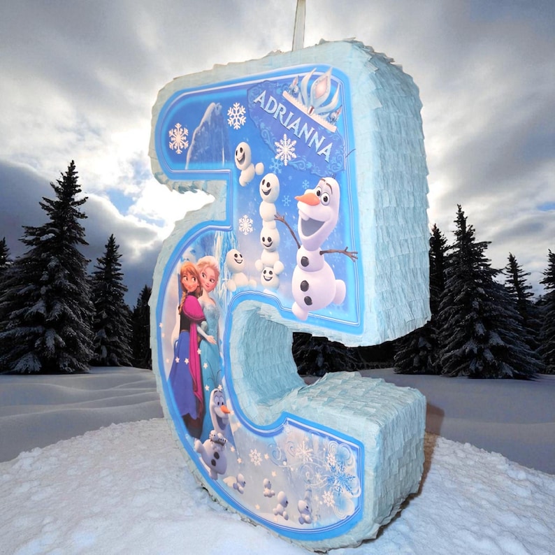 Puede incluir: Una pi&ntilde;ata grande con el n&uacute;mero 5 de color azul claro con tem&aacute;tica de Frozen. La pi&ntilde;ata presenta im&aacute;genes de Elsa, Anna y Olaf, junto con copos de nieve y el nombre "Adrianna". Los bordes est&aacute;n deshilachados y el fondo muestra un paisaje nevado.