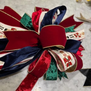 May include: A large, decorative Christmas bow featuring a combination of red, navy blue, and green velvet ribbons. Some ribbons have a gold trim and a pattern of toy soldiers. The bow is a festive holiday decoration.