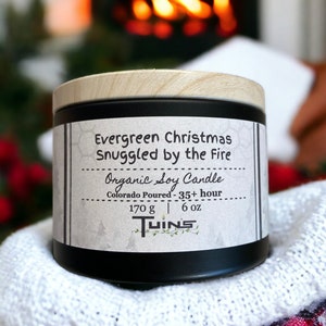 May include: A black candle jar with a wooden lid. The label reads "Evergreen Christmas Snuggled by the Fire Organic Soy Candle Colorado Poured - 35+ hour 170 g | 6 oz TWINS".