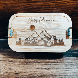 Könnte beinhalten: Eine Holzlunchbox mit einer Bergszene und dem Text "Gipfelstürmer" auf dem Deckel graviert. Die Lunchbox hat zwei Metallverschlüsse.