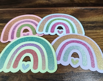 May include: Four rainbow-shaped paper cutouts with glitter accents. Each rainbow has a small heart in the center. The rainbows are in shades of pink, blue, green, and purple.