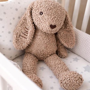 Könnte beinhalten: Ein braunes Plüsch-Hasen-Spielzeug mit langen Ohren und einer herzförmigen Nase sitzt in einem weißen Kinderbett. Der Hase hat ein strukturiertes, flauschiges Aussehen und das Wort "Theo" ist auf einem Ohr gestickt. Das Kinderbett hat eine sternförmige Bettumrandung.