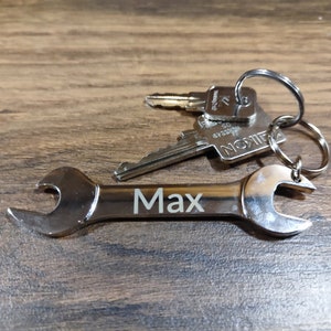 Puede incluir: Un llavero de llave inglesa plateado con el nombre "Max" grabado en él. El llavero está unido a un juego de llaves con un anillo plateado.