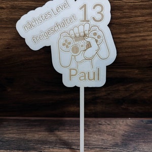 Könnte beinhalten: Ein weißes Holzschild mit einem braunen Holzstab, der an der Unterseite befestigt ist. Das Schild zeigt einen braunen Videospielcontroller mit einer Hand, die ihn hält. Das Schild hat auch den Text "nächstes Level freigeschaltet" und die Zahl "13" in braun. Das Schild hat auch den Namen "Paul" in braun.