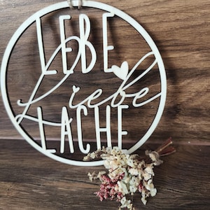 Könnte beinhalten: Ein rundes Holzschild mit dem deutschen Schriftzug "Lebe Liebe Lache" in dekorativer Schrift. Unter dem Schild liegt ein kleines Bündel Trockenblumen. Das Schild wird an einer natürlichen Schnur aufgehängt.