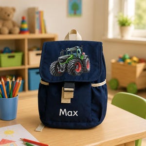 Könnte beinhalten: Ein dunkelblauer Rucksack mit einer Grafik eines grünen Traktors auf der oberen Klappe und dem Namen Max in weißen Buchstaben auf der Vordertasche. Der Rucksack hat einen beigen Tragegriff, beige Kanten und einen schwarzen Kunststoff-Schnallenverschluss.