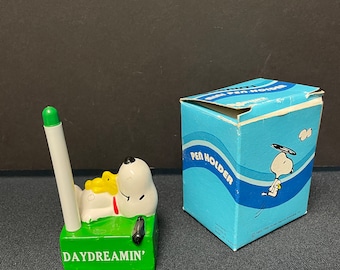 May include: A Snoopy pen holder with a green base that reads "DAYDREAMIN'". The pen holder features Snoopy and Woodstock. A white pen with a green tip is in the holder. A blue box with the words "PEN HOLDER" is next to the pen holder.