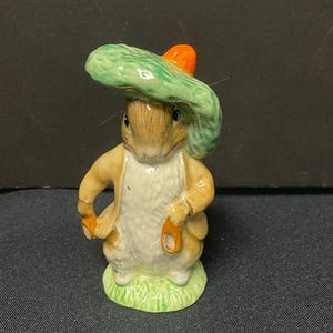 May include: A small ceramic figurine of a rabbit wearing a green hat with an orange brim. The rabbit is standing on a green base.