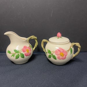 May include: A vintage cream-colored sugar and creamer set with a pink rose design. The set has green leaves and stems and a white background.