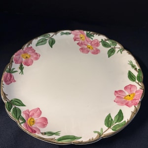 May include: A white ceramic plate with a scalloped edge, adorned with pink rose blossoms, green leaves, and a gold-colored trim. The floral design is hand-painted, creating a vintage aesthetic. The plate is suitable for serving food.