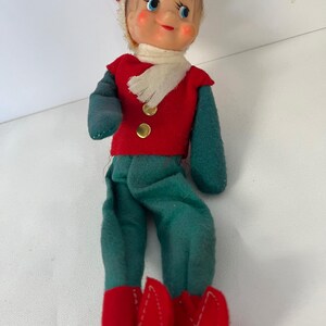 May include: A vintage Christmas elf doll with a painted face, wearing a red vest with gold buttons, green pants, and red boots. The doll has a white scarf and a hat with a red and white stripe.
