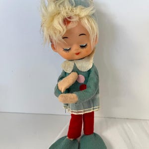 May include: A vintage doll with a teal outfit, red pants, and large teal shoes. The doll has blonde hair, a teal hat, and a white collar. The doll's eyes are closed, and it has a red mouth.