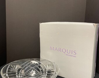 May include: A clear crystal Marquis by Waterford bowl with a decorative cut glass design. The bowl has a wide, oval shape and sits on a small, round base. A white box with the brand name is behind the bowl.