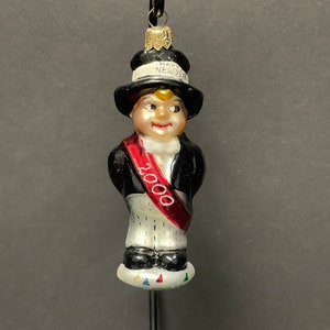 May include: A glass ornament depicting a person wearing a black top hat with the text "Happy New Year" on it. The person is wearing a red sash with the year "2000" on it.