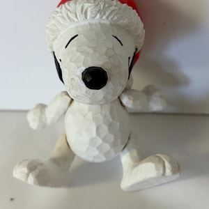 May include: A white Snoopy figurine with a textured, carved wood appearance, wearing a red Santa hat with a white brim. The character has a black nose, closed eyes, and is depicted in a standing pose with arms outstretched to the sides.