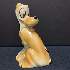 May include: A ceramic figurine of a cartoon dog, Pluto, from Disney. The figurine is brown and white with black accents. Pluto is sitting with his head turned to the side and his mouth open in a smile.